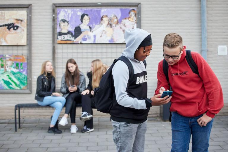 Decoratieve foto van twee leerlingen op een schoolplein. Een van de leerlingen houdt een smartphone vast terwijl ze er samen naar kijken.