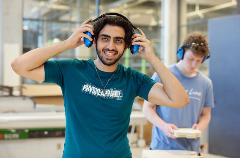 Decoratieve foto van een mbo-student die naar de camera lacht en zijn gehoorbescherming opdoet. Op de achtergrond werkt een medestudent aan een werkbank, ook met gehoorbescherming op.